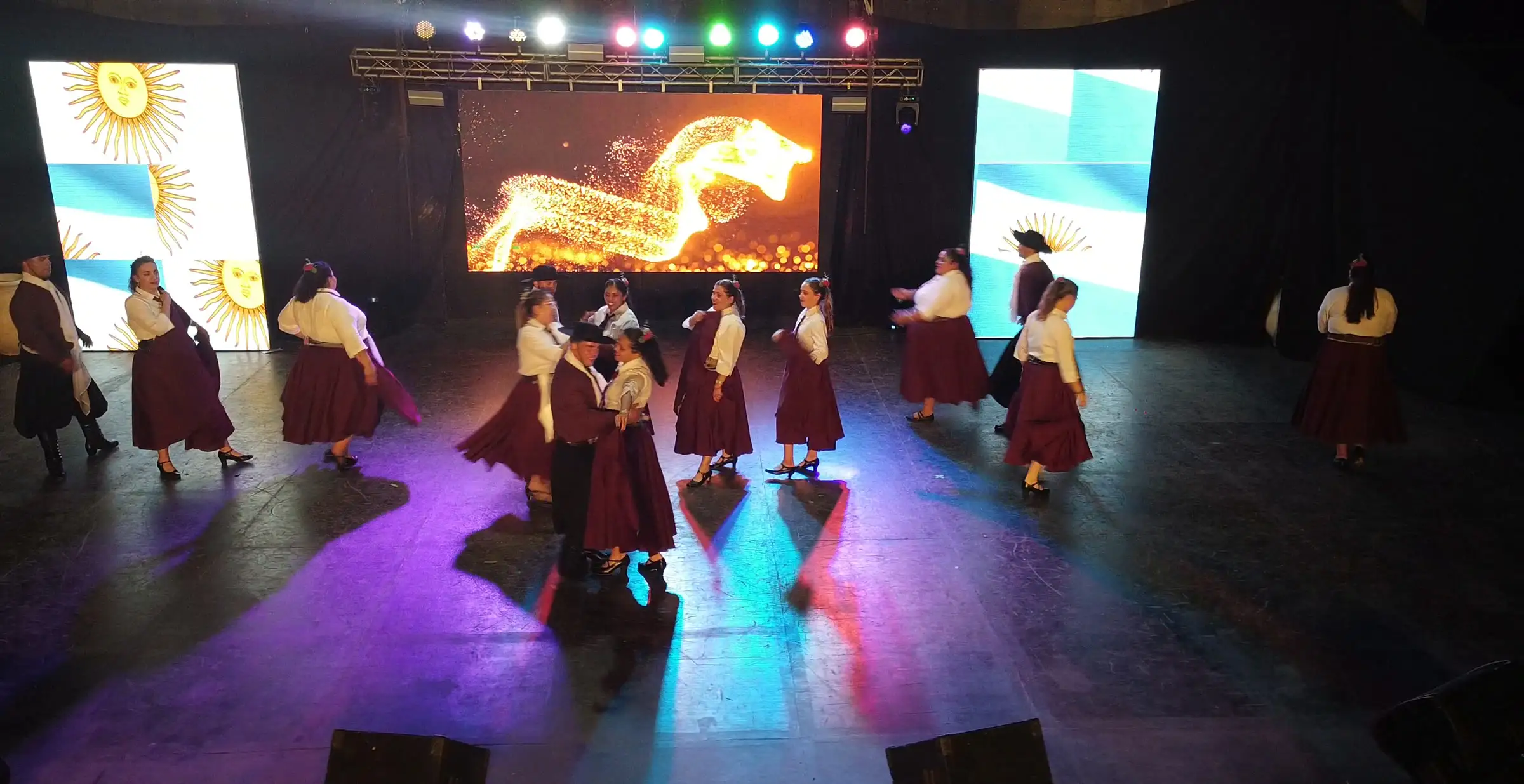 Vista aérea de un escenario con pantallas LED, truss de luces encendido y un ballet folklórico bailando.